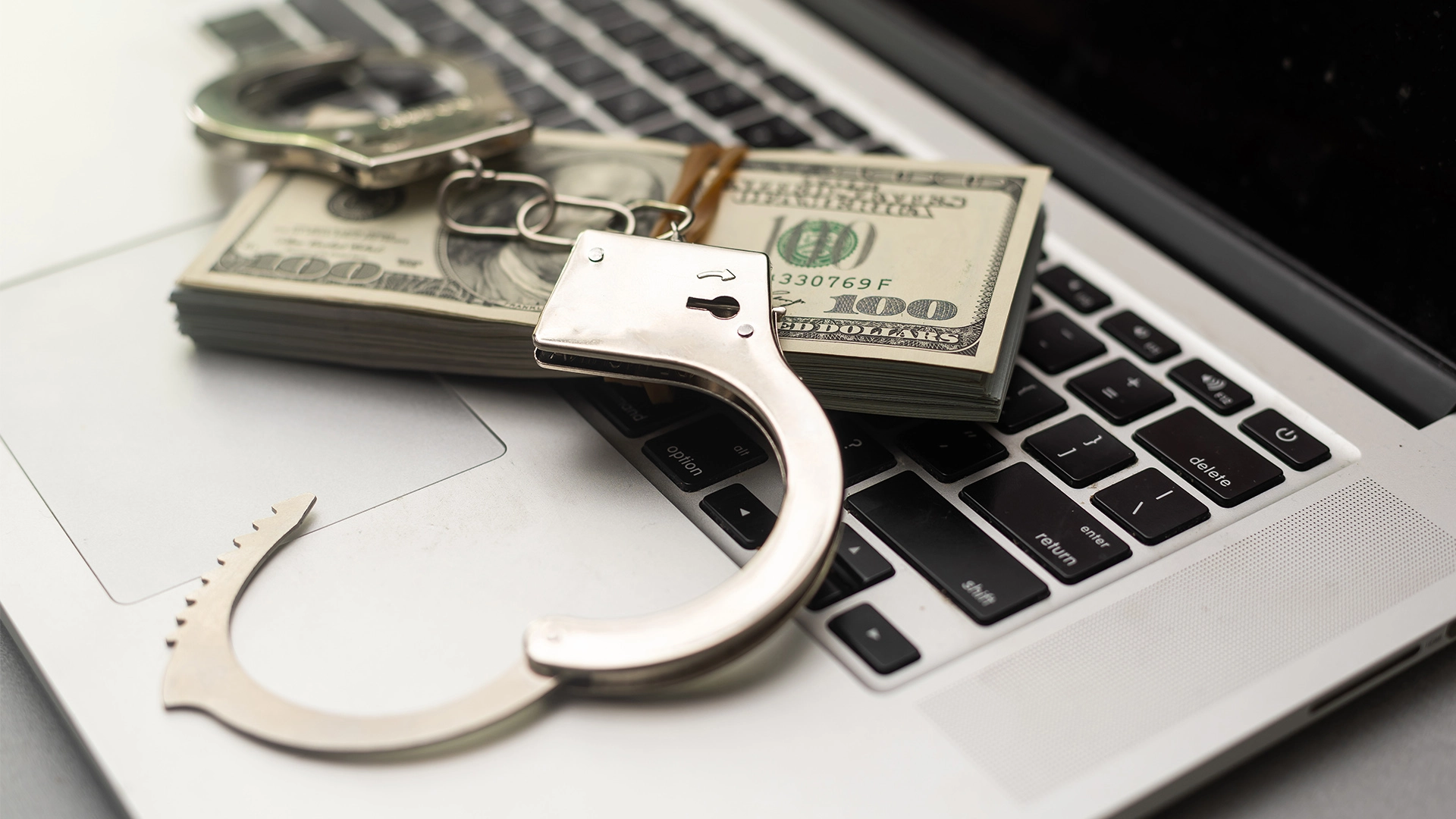 A pair of handcuffs and cash resting on a laptop keyboard representing white collar crime